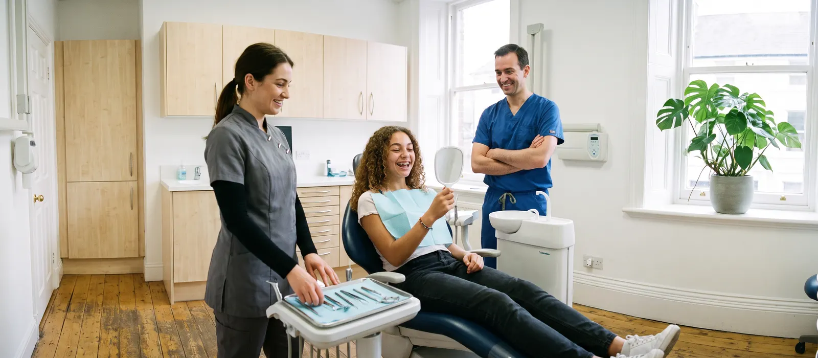 A patient checks new braces in a hand mirror while the orthodontist and assistant stand beside the chair